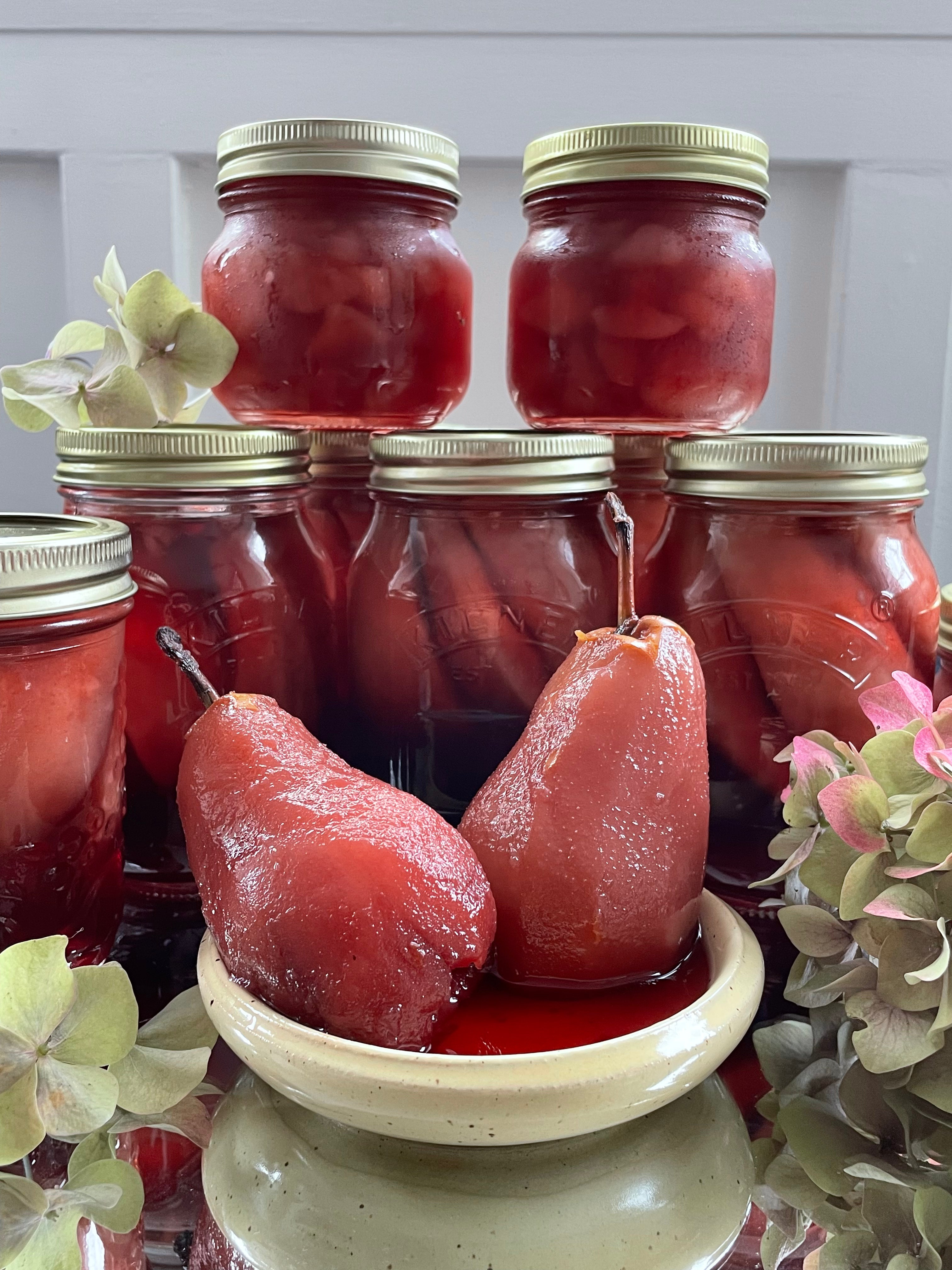 Jars of poached pears with a decorative bowl containing two poached pears on a white surface.